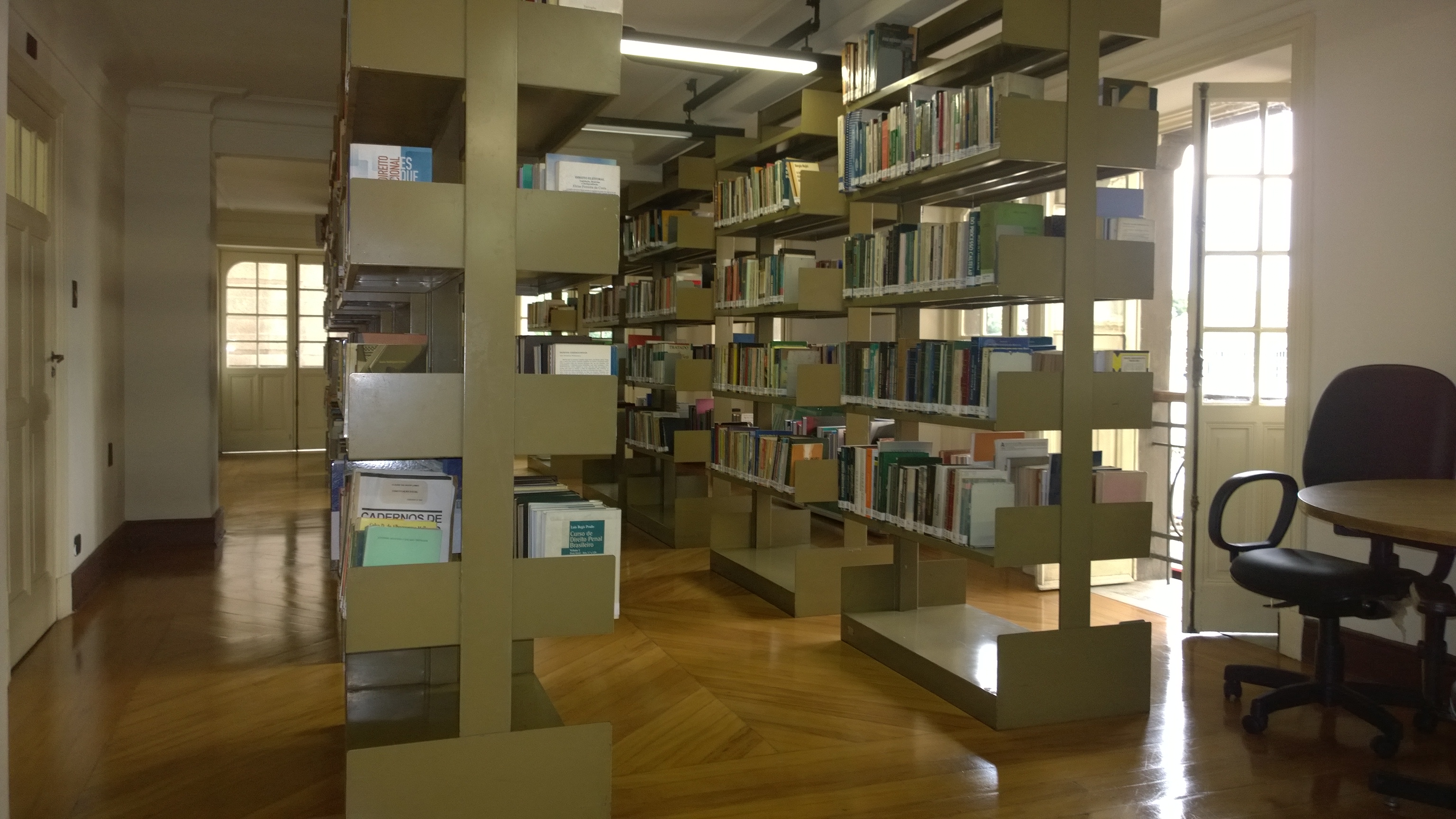 A imagem contém prateleiras de livros no interior da sala de Biblioteca. Uma fileira delas à esquerda e outra à direita, ao lado de uma cadeira vinho. A sala está iluminada pela luz externa, janelas encontram-se ao redor da sala. A biblioteca é localizada no Centro de Estudos Jurídicos.