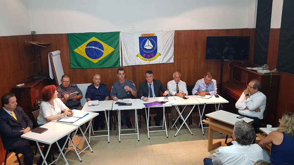 Onze pessoas sentadas em auditório, posicionadas em meio círculo, bandeiras do Brasil e Conseg ao fundo