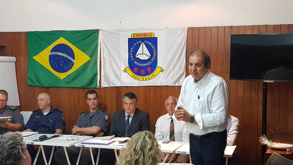 Subprefeito em pé, oito pessoas sentadas e bandeiras do Brasil e do Conseg ao fundo