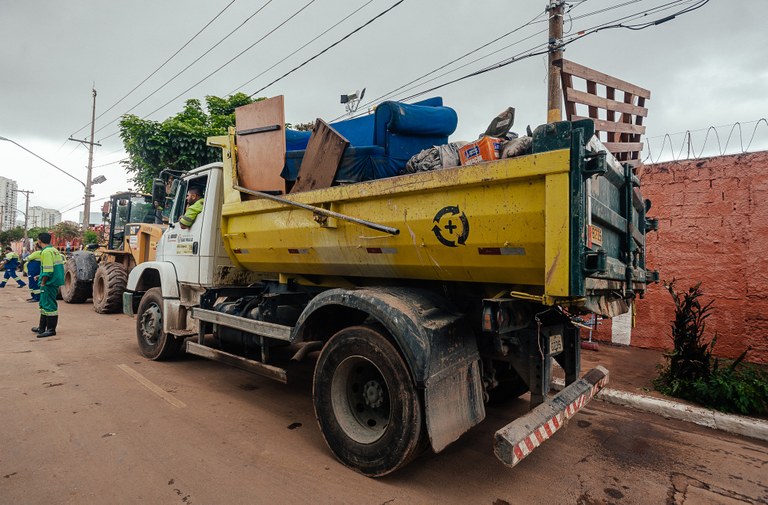 Caminhão na cor amarela transportando móveis, concreto e madeira. Do lado esquerdo é possível ver três funcionários da Subprefeitura Ipiranga, vestidos de uniformes na cor verde, recolhendo o entulho