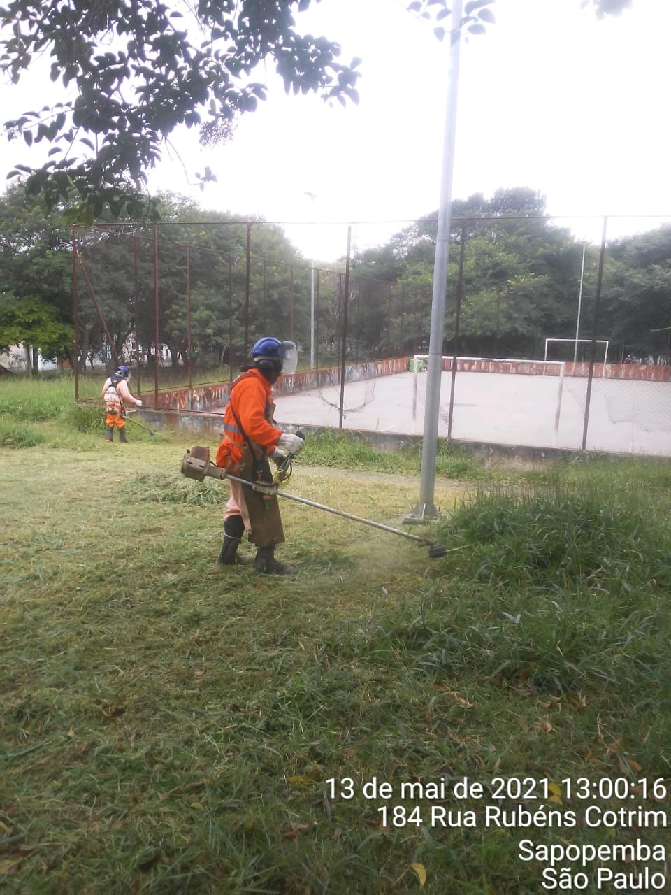 Trabalhador com uniforme laranja, roça grama ao lado de uma quadra de esportes, em uma praça. 