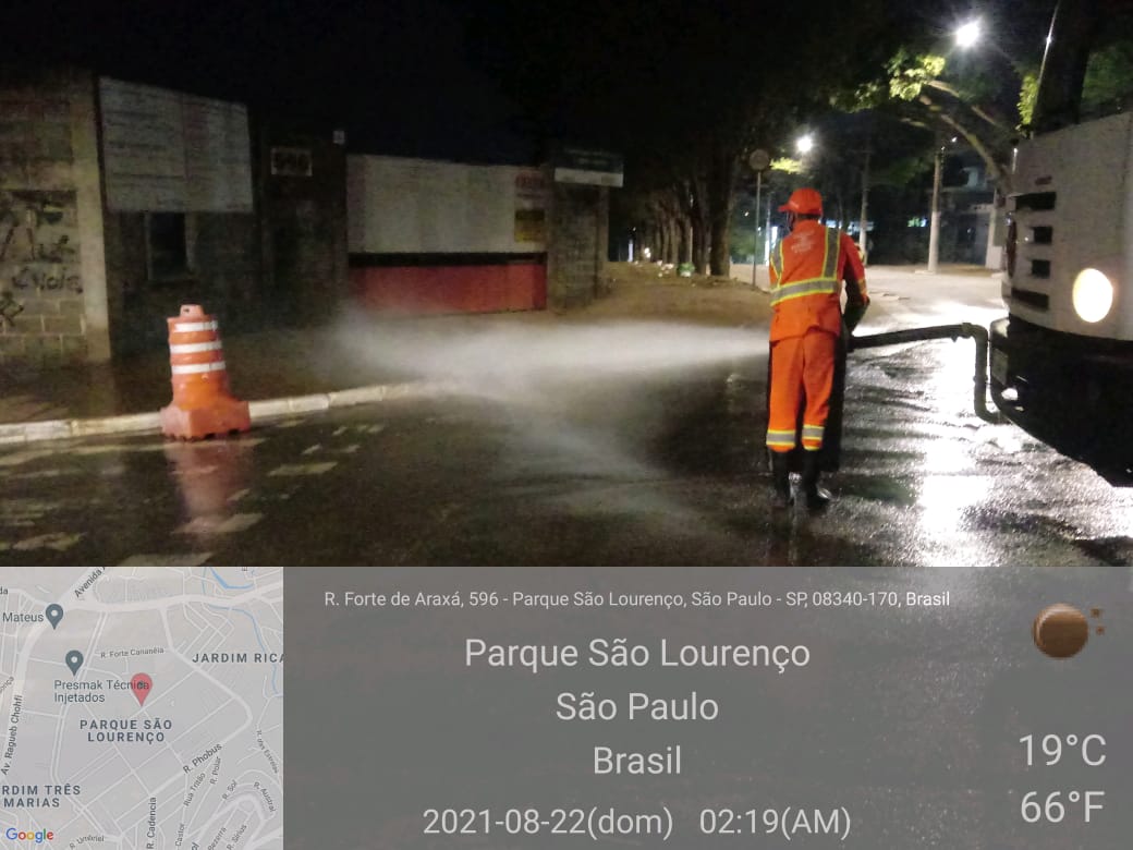 Homem com uniforme laranja segura mangueira com esguicho de água, em uma rua, na frente de um caminhão, com farol aceso e sinalização com cone laranja na pista. 