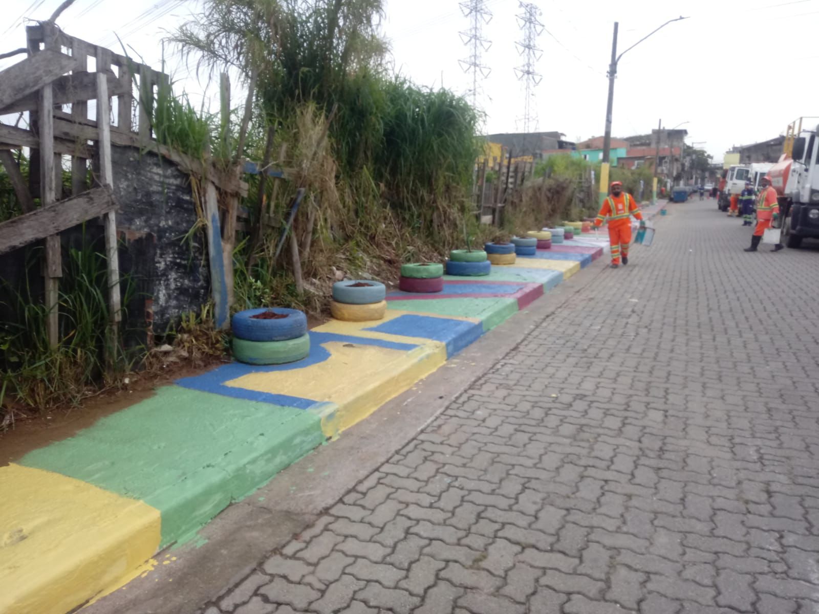 Pneus coloridos, em blocos de dois, enfileirados em uma calçada, também pintada de amarelo, verde, azul e magenta. Nos pneus foi colocada terra . m