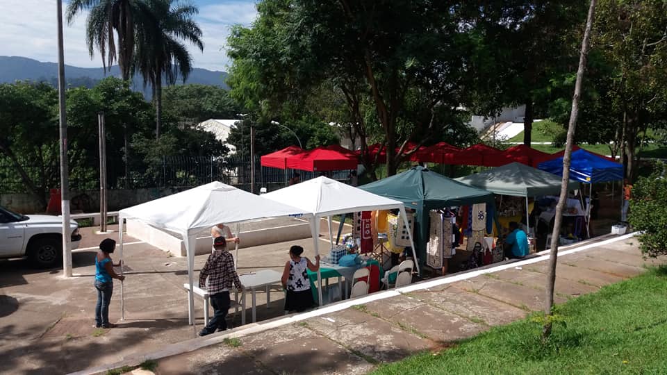 Foto das Tendas da Feira de Artesanato do Evento, há tentas nas cores branco, azul, verde e vermelho, há quatro pessoas montando uma tenda branca, ao fundo há vegetação.