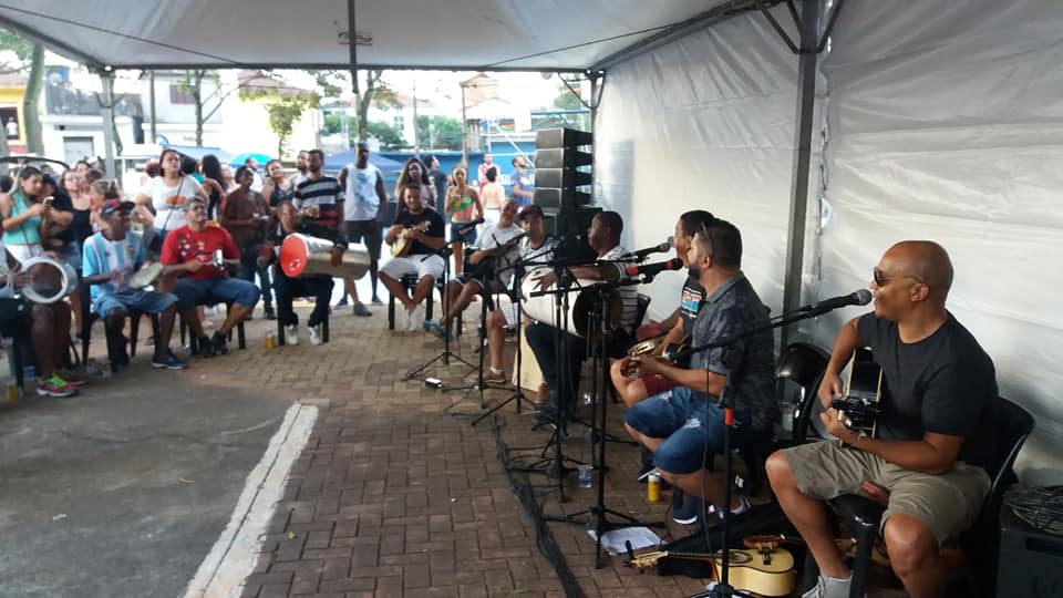 Foto do "Grupo Fim De Semana", se apresentando na praça, há cerca de dez músicos, eles estão cantando e tocando instrumentos, dentre os instrumentos há violão,  batuque, pandeiro, e outros, ao fundo estão os munícipes assistindo a apresentação.