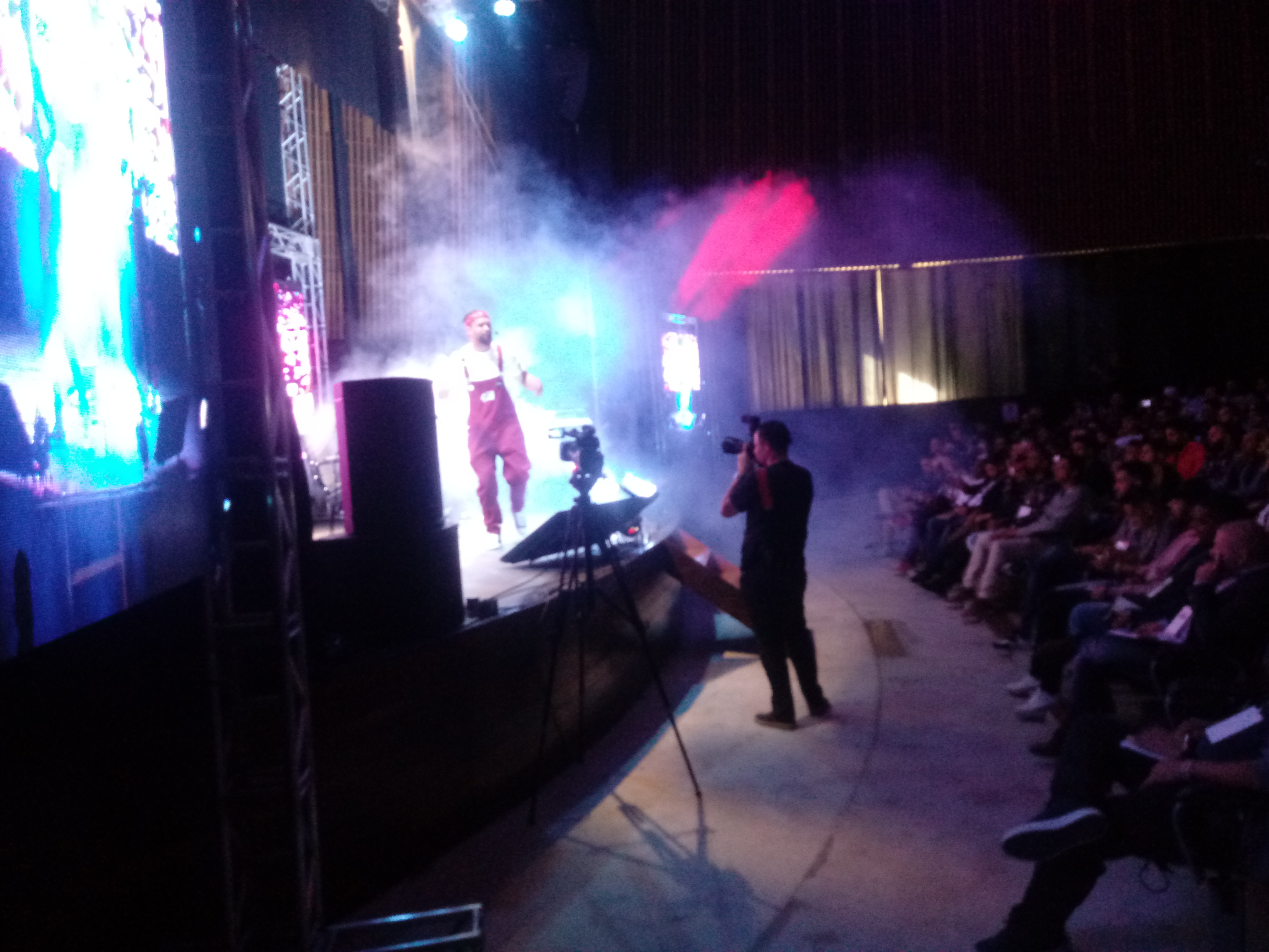 Artista se apresenta em palco, com efeito visual de gelo seco. Veste macacão vermelho, camiseta branca e usa bandana vermelha.