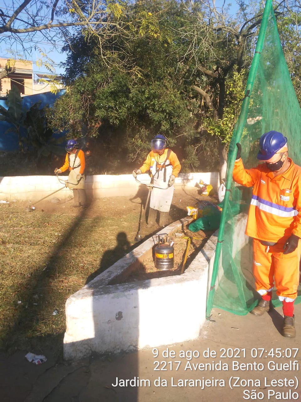 Cinco trabalhadores com uniforme laranja trabalham na conclusão de um sarjetão, que já está concretado. 