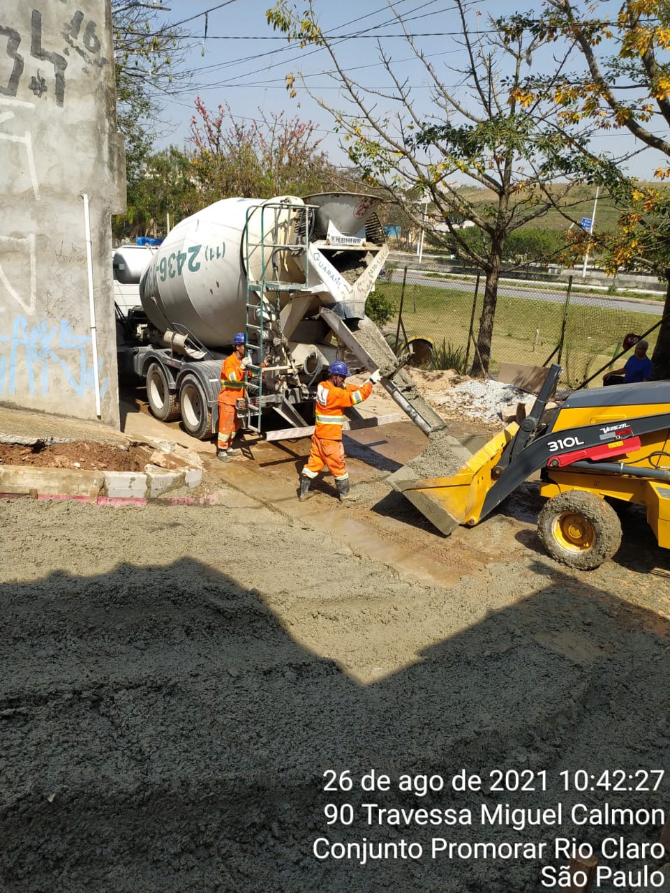 Um caminhão betoneira está parado, com a bica de concreto estendida, para a caçamba de uma pá carregadeira. Dois homens de uniforme laranja estão em pé entre os dois equipamentos. 