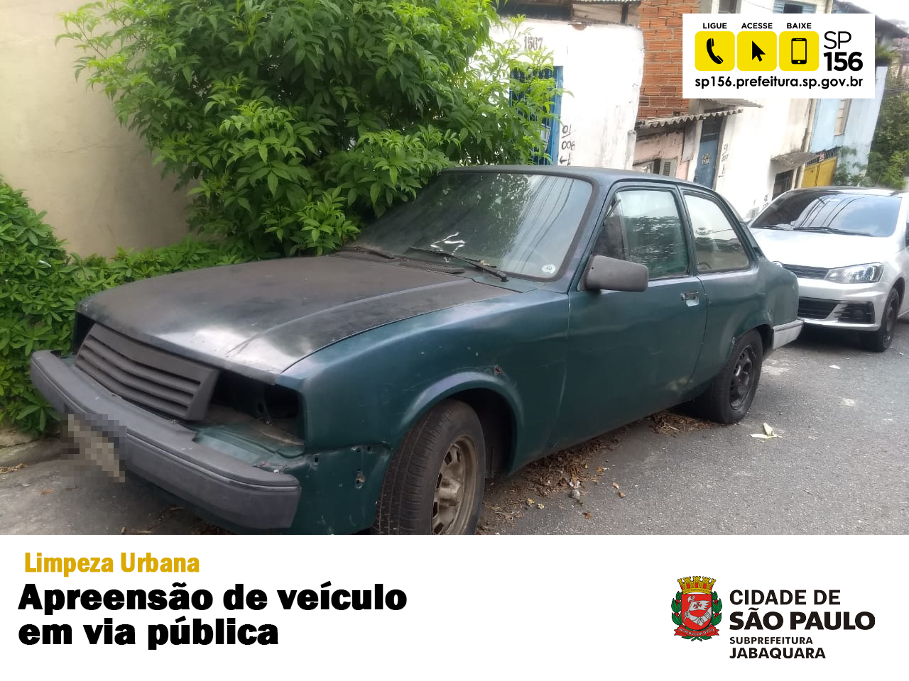 Carro abandonado perto de uma árvore, a fotografia contém uma faixa brancano rodapé escrito "Limpeza Urbana - Apreensão de veículos em via pública" e mais ao canto inferior direito o logotipo da Subprefeitura Jabaquara. No canto superior direito tem o ícone com o telefone do 156.