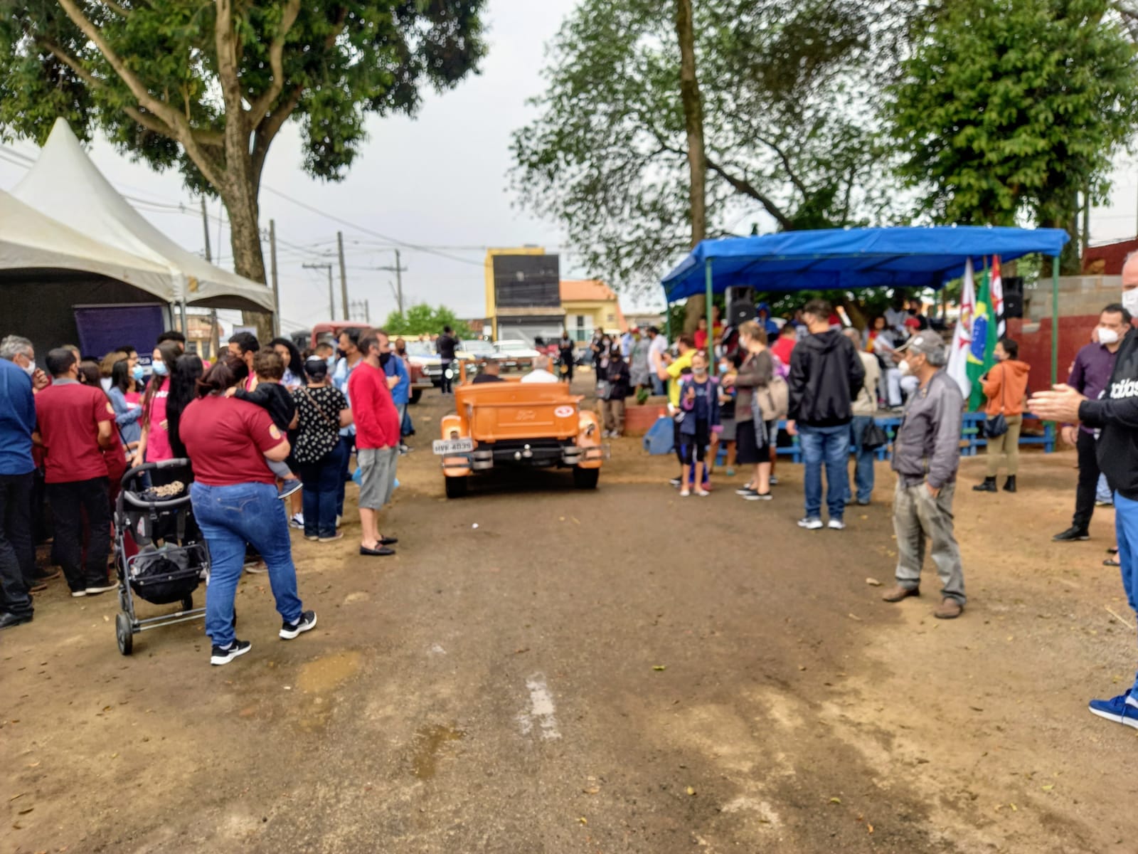 Pessoas enfileiradas à direita e à esquerda, admiram a passagem de uma pequena caminhonete antiga, pintada de laranja.