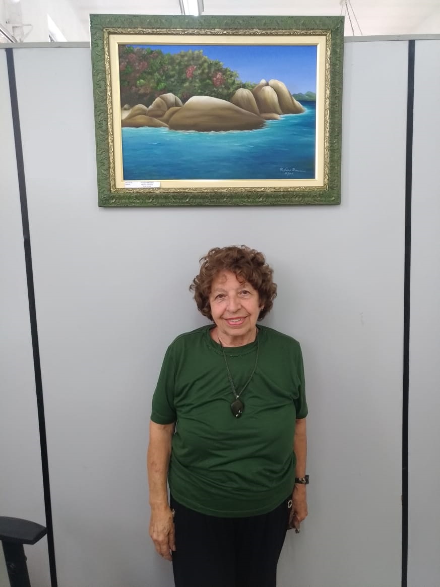 Imagem colorida, com a artista plástica Rosa Branchini vestindo camiseta verde, calça preta, colar grande e cabelo curto, em cima um quadro de uma paisagem pintado por ela.