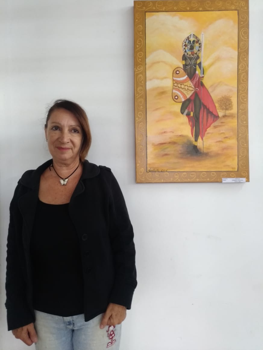 Imagem colorida, da artista plástica Rosália Gonsalves, camiseta e blazer pretos, colar de borboleta, no seu lado direito o quadro pintado por ela.
