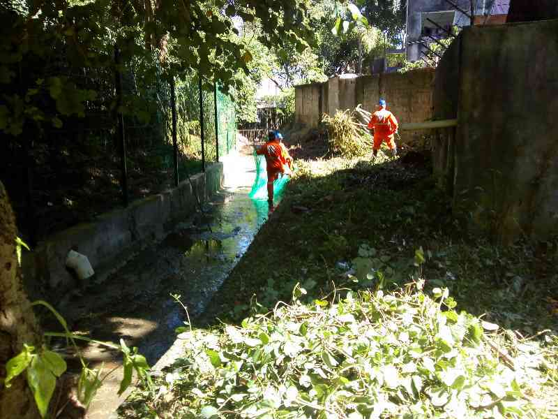 Dois funcionários da Subprefeitura Ipiranga vestidos com uniformes na cor laranja realizando a limpeza na margem do córrego São José. Envolta do córrego é possível ver algumas árvores.