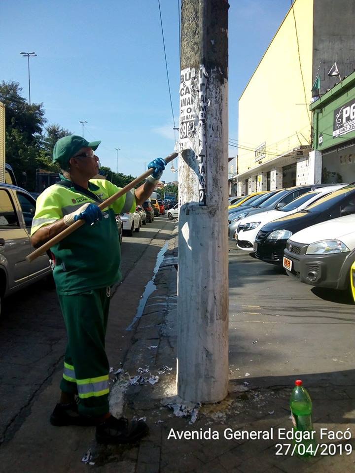 Funcionário devidamente uniformizado (uniforme em tons de verde claro e escuro) realizando a rapasgem de propaganda irregular de um poste