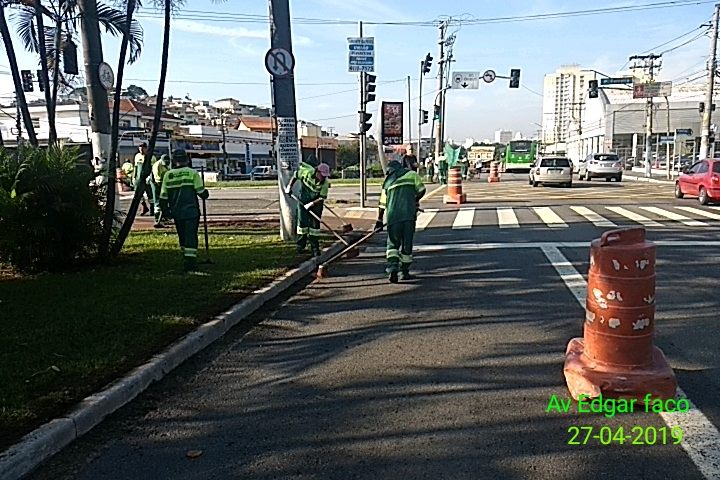 Equipe de funcionários de limpeza devidamente uniformizados (uniforme em tons de verde) realizando o serviço de varrição, bem ao fundo outra equipe realizando o serviço de corte de mato.