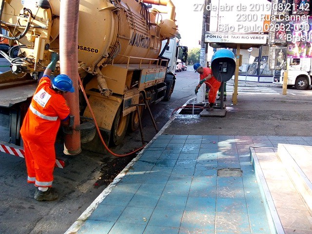 Dois funcionários devidamente uniformizados (Unifome de cor laranja) realizando a limpeza mecânicada de bueiro 