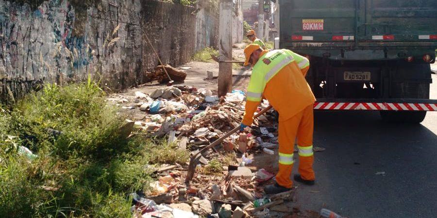 Funcionário da prefeitura retirando entulho de calçada com enxada; atrás dele, outro funcionário e um caminhão.