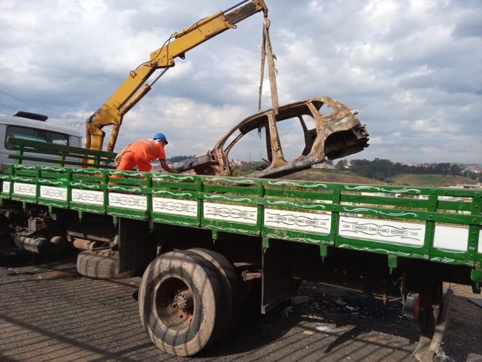 Carcaça de um carro queimado é removida, com guincho para a carroceria de um caminhão. Um ajudante ajeita a parte dianteira no assoalho.