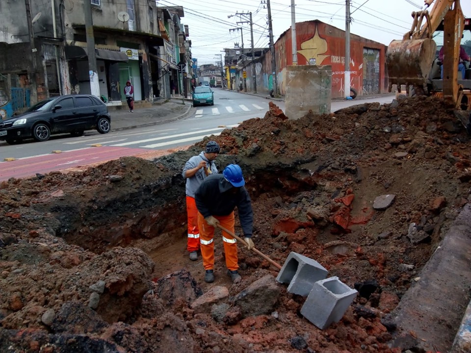 Dois homens vestidos de uniforme laranja, da Prefeitura, trabalham dentro de um buraco aberto com máquina.