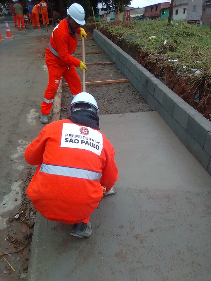 Dois trabalhadores com uniforme laranja da Prefeitura trabalham na construção de uma calçada.