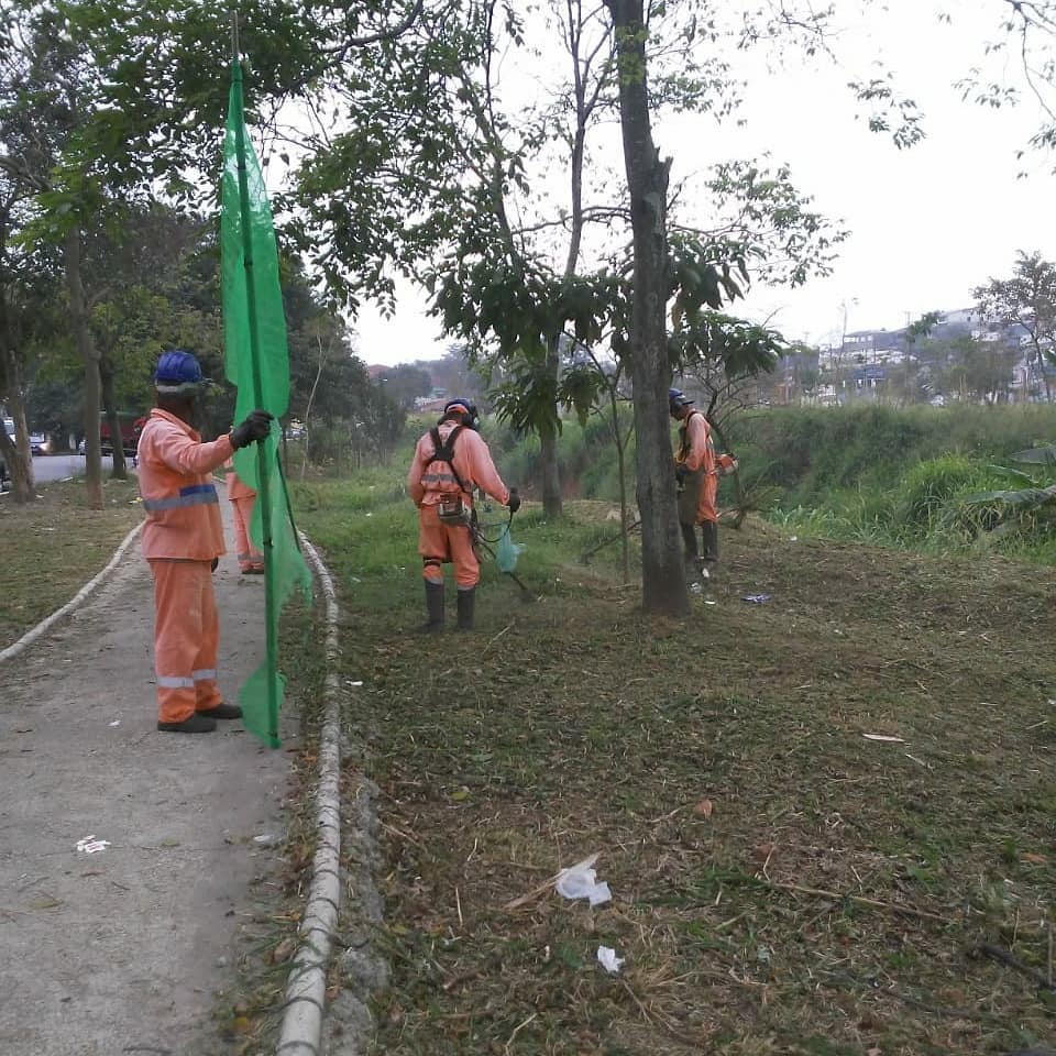 Dois funcionários cortam a grama com roçadeiras; outros dois seguram tela de proteção.