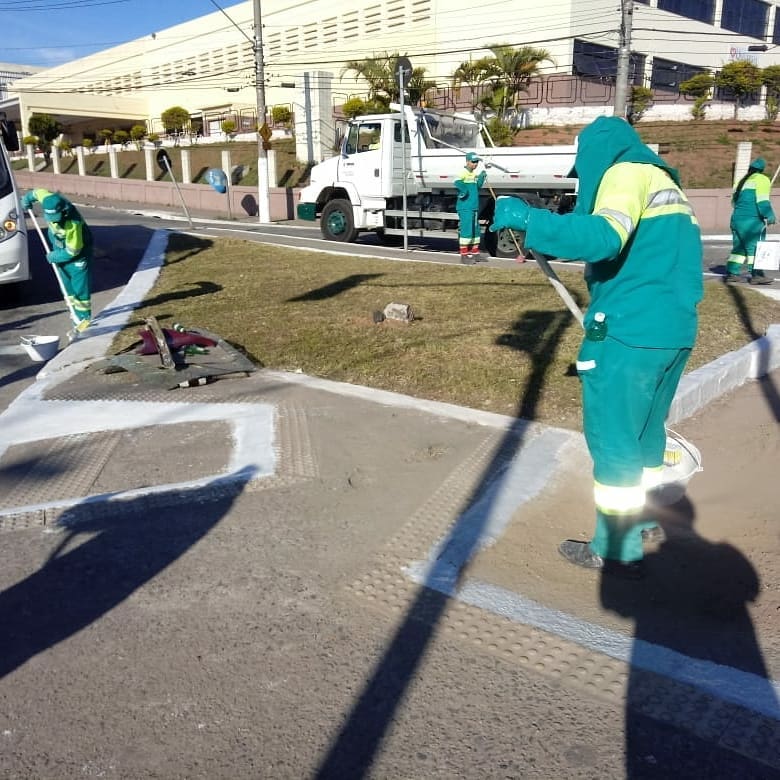 Funcionários varrem a calçada uniformizados e com proteção na cabeça, devido ao forte sol; um caminhão de lixo está ao fundo da imagem.