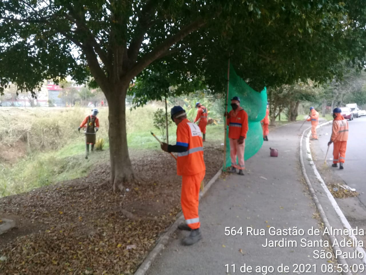 Funcionários da Prefeitura, com macacão laranja, fazem poda de grama na praça da foto identificada como rua Gatão de Almeida, Jardim Santa Adélia