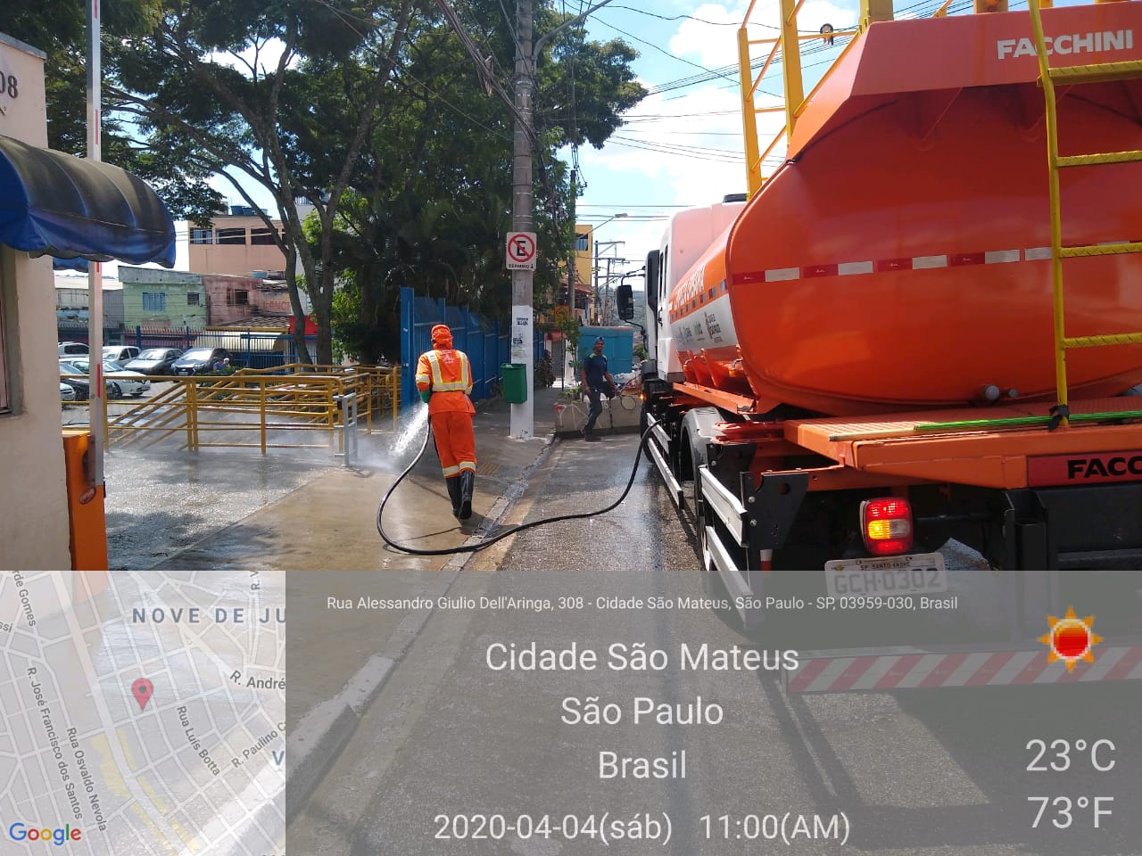 Caminhão com tanque laranja está parado e trabalhador lança jatos de água na calçada.