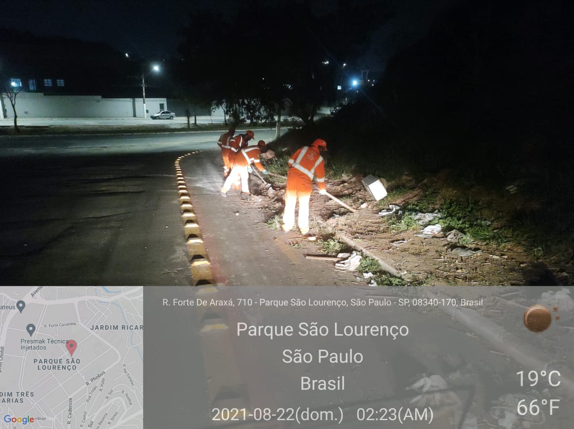 Com reflexos das luzes da rua, dois homens com uniforme laranja recolhem sujeira na calçada, usando enxada. 