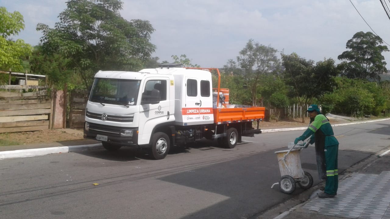 Caminhão de limpeza está parado em frente uma área verde, em uma rua vazia. Funcionária está com carrinho de tinta para pintura de guias.