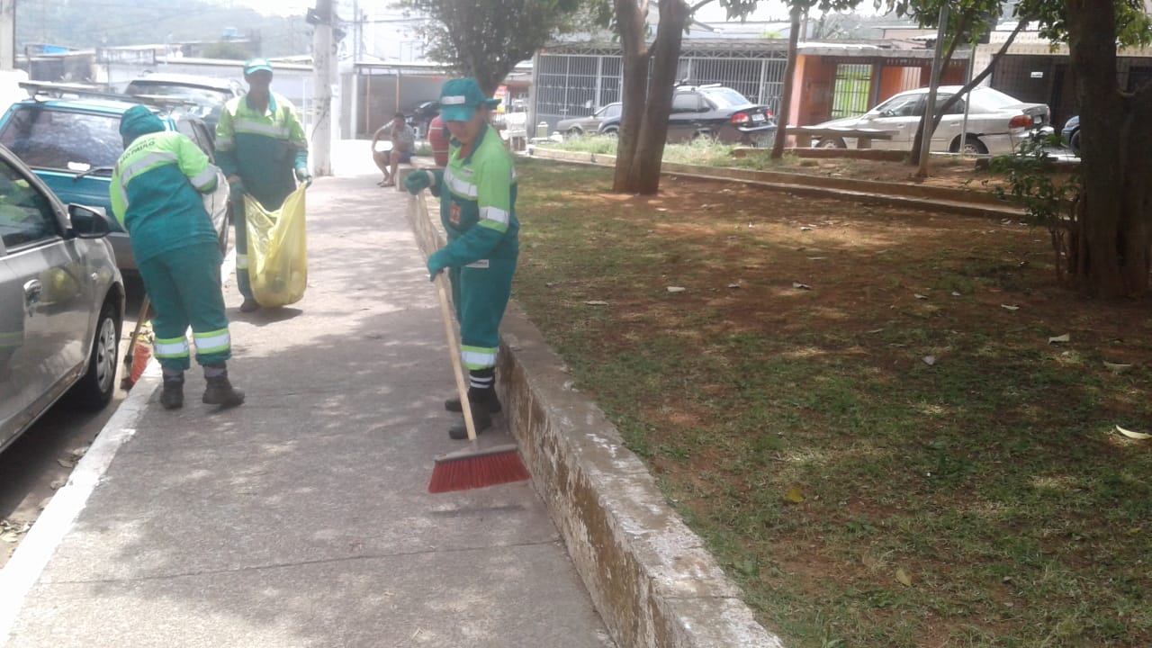 Trabalhadores com uniforme verde recolhem lixo em uma praça, um deles segura saco plástico amarelo, já com uma certa quantidade.