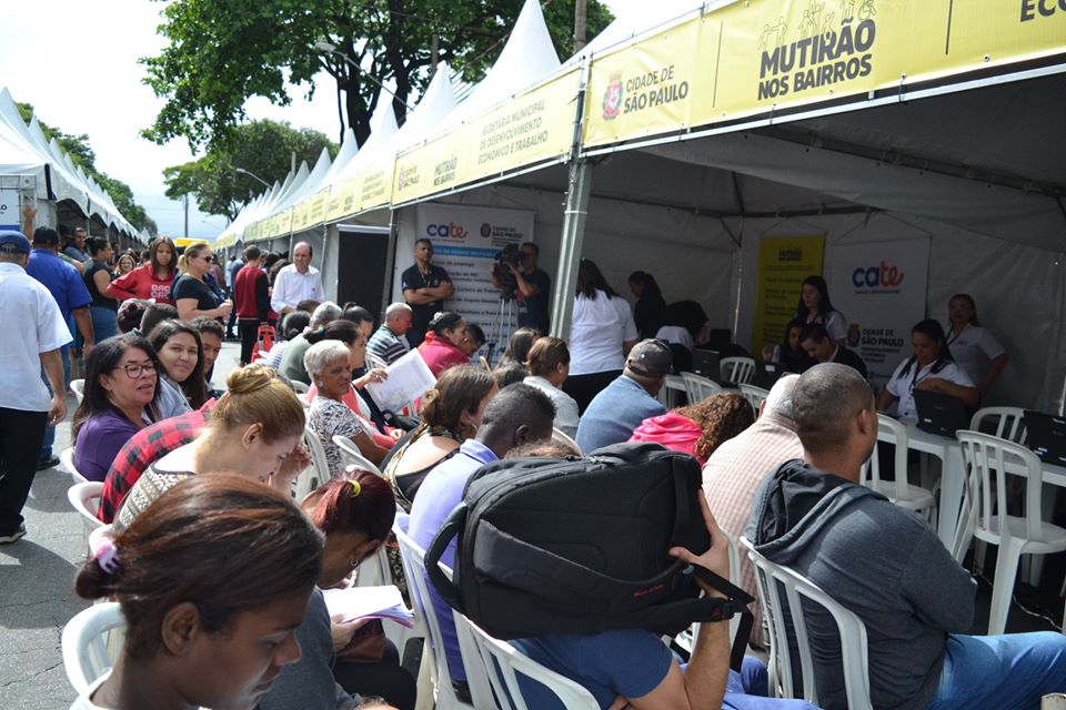 Um grupo de pessoas está sentado sobre cadeiras brancas em frente à tenda do Centro de Apoio ao Trabalho e Empreendedorismo (CATe), enquanto esperam pelo atendimento.  