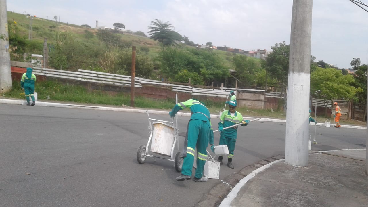 Cinco funcionários com uniforme verde e um de uniforme laranja fazem a pintura de guias  e postes em uma rua