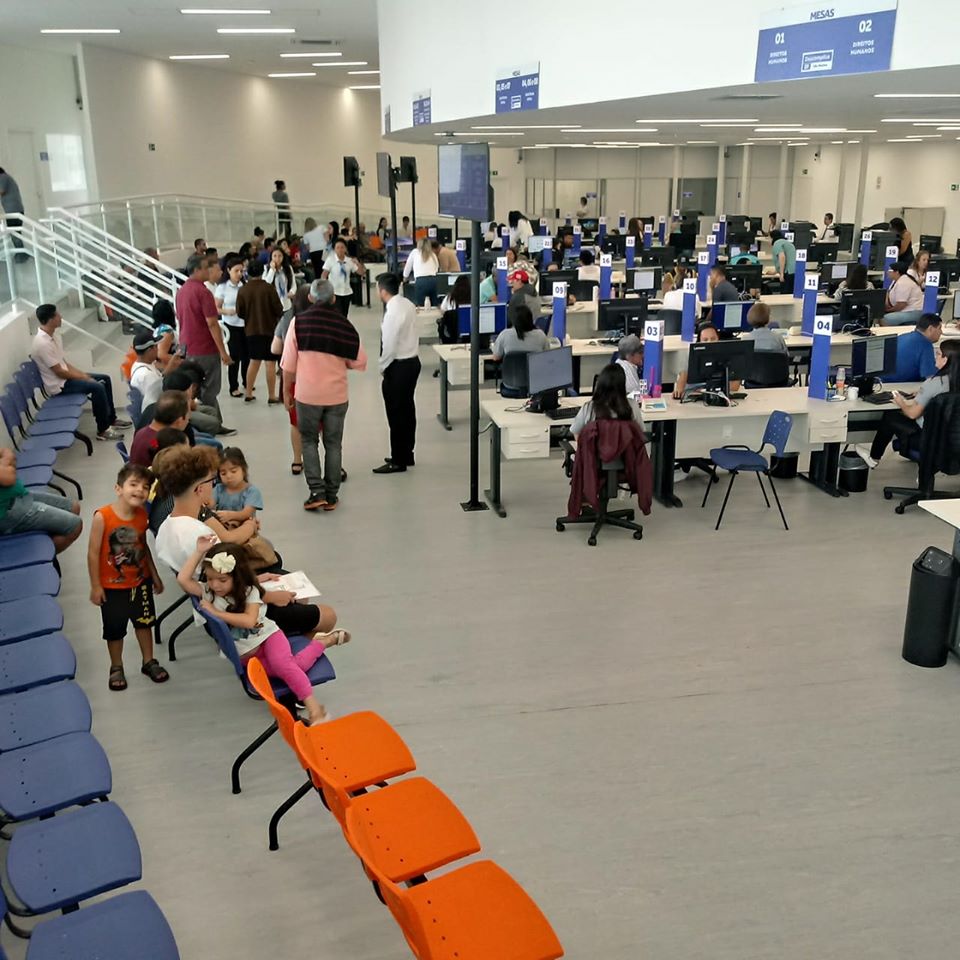 Salão com piso branco, cadeiras azuis e uma parte em laranja, indicando preferencial. Mesas de atendimento, com computadores. Pessoas aguardam ser chamadas e outras estão sendo atendidas.