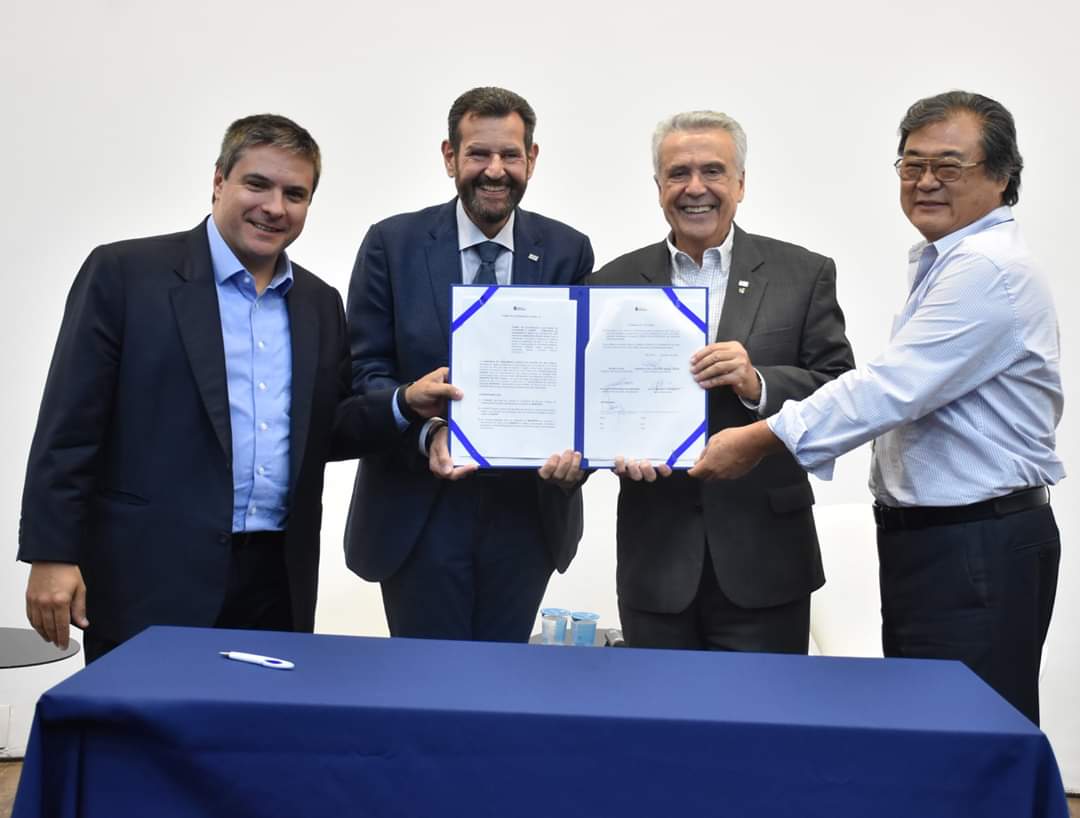A imagem mostra quatro homens adultos em pé, sorrindo e segurando documentos. Eles estão posicionados atrás de uma mesa coberta com uma toalha azul escura.  O homem mais à esquerda tem cabelos castanhos claros e usa um terno escuro com uma camisa azul clara. Ele está olhando para a câmera e sorrindo.  O segundo homem da esquerda, no centro da imagem, tem barba e usa um terno escuro com uma gravata azul escura. Ele está segurando um documento com as duas mãos, mostrando-o para a câmera, e também está sorrindo.  O terceiro homem da esquerda, ao lado do anterior, tem cabelos grisalhos e usa um terno escuro com uma camisa branca. Ele também está segurando um documento e sorrindo para a câmera.  O quarto homem, mais à direita, tem cabelos escuros e óculos. Ele veste uma camisa clara e está segurando uma das bordas do documento que os outros dois homens no centro estão mostrando. Ele também está sorrindo.  Todos os homens parecem estar em um ambiente interno, com uma parede branca ao fundo. Há alguns objetos sobre a mesa, mas não estão claramente definidos. A iluminação parece ser artificial.  A cena sugere um momento de assinatura de um acordo ou algum tipo de formalização, dada a presença dos documentos e a expressão de satisfação dos homens.