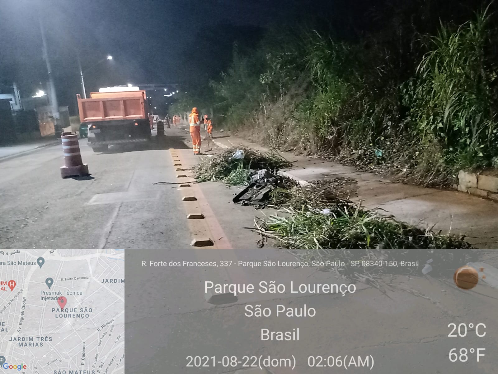 Montes de lixo e detritos estão depositados na beira do asfalto. à frente, dois homens de uniforme laranja trabalham com ferramentas na mão e ao fundo, um caminhão basculhante está parado. 