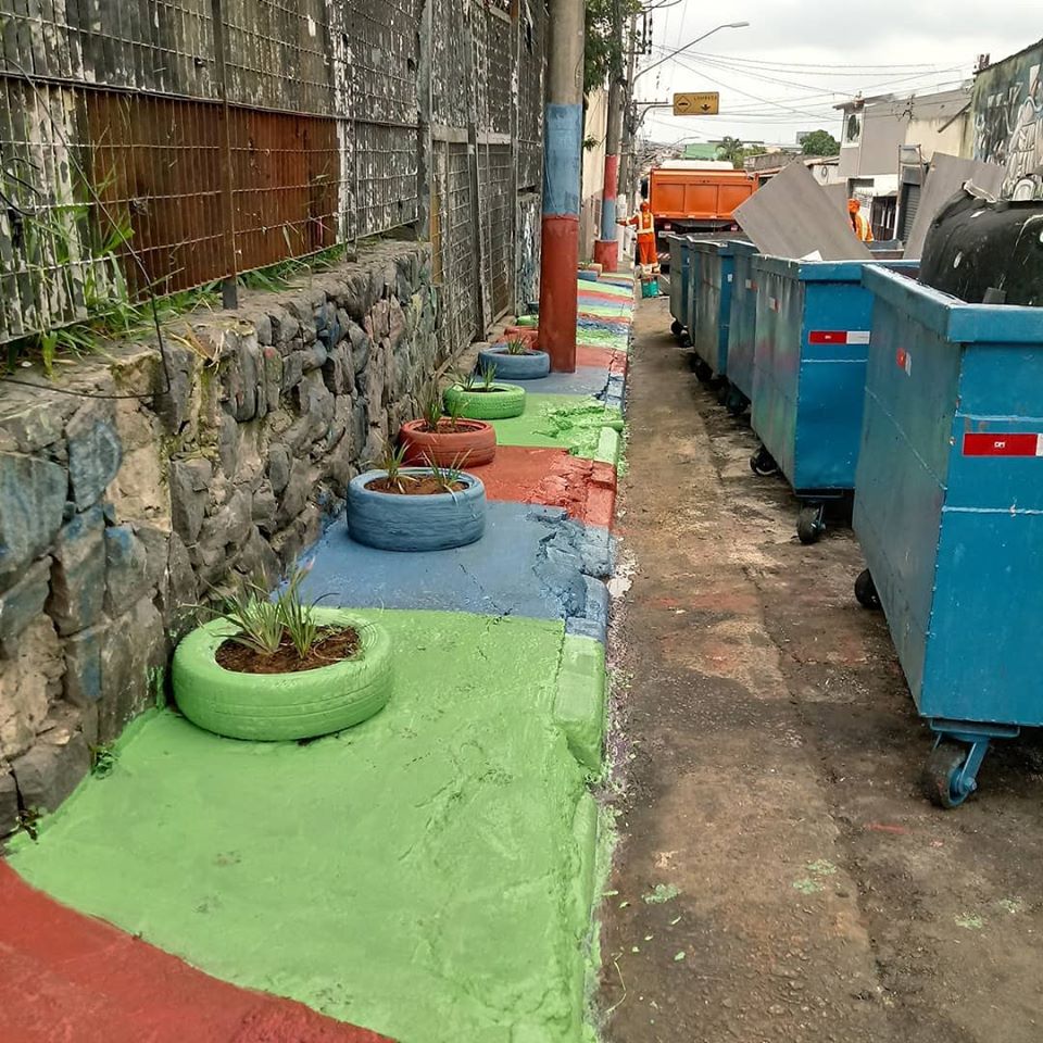 à esquerda, calçada recém reformada, com pintura nas cores vermelho, verde e azul, além de pneus reutilizados como vaso para plantas. à direita, caçambas cheias de entulho retirados do local.