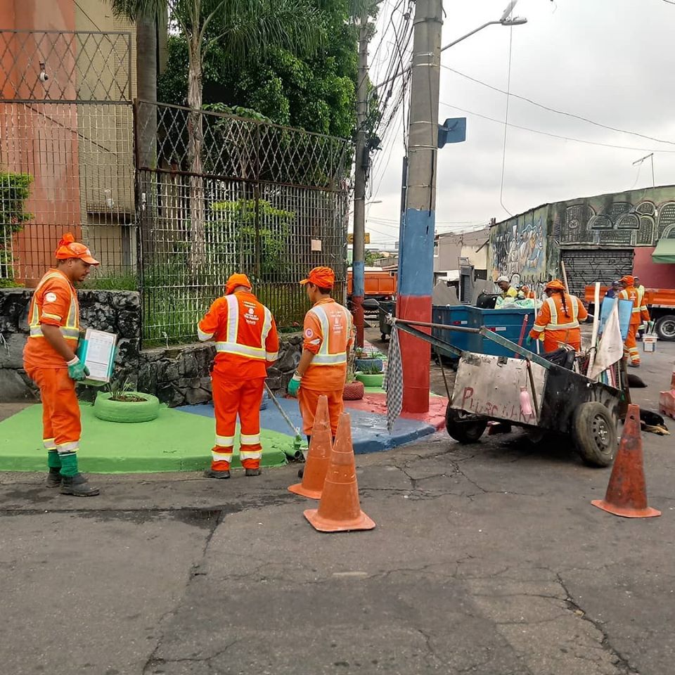 Funcionários recolhem lixo e pintam calçada em cruzamento de ruas; local está isolado por cones e à direita há um carrinho de mão.