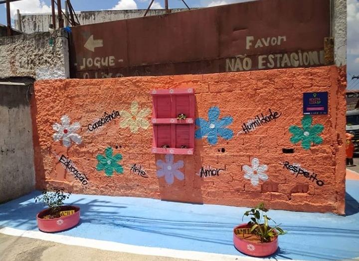 Imagem do muro pintado de laranja e com pinturas de flores, a calçada esta pintada da mesma cor, existem pneu pintados de azul que estão sendo usados como vasos para flores