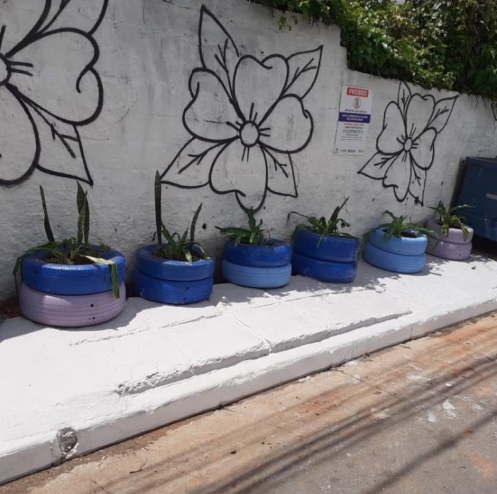 Imagem de um muro com pintura de flores com pneu pintados de azul encostados no muro com muda de flores dentro