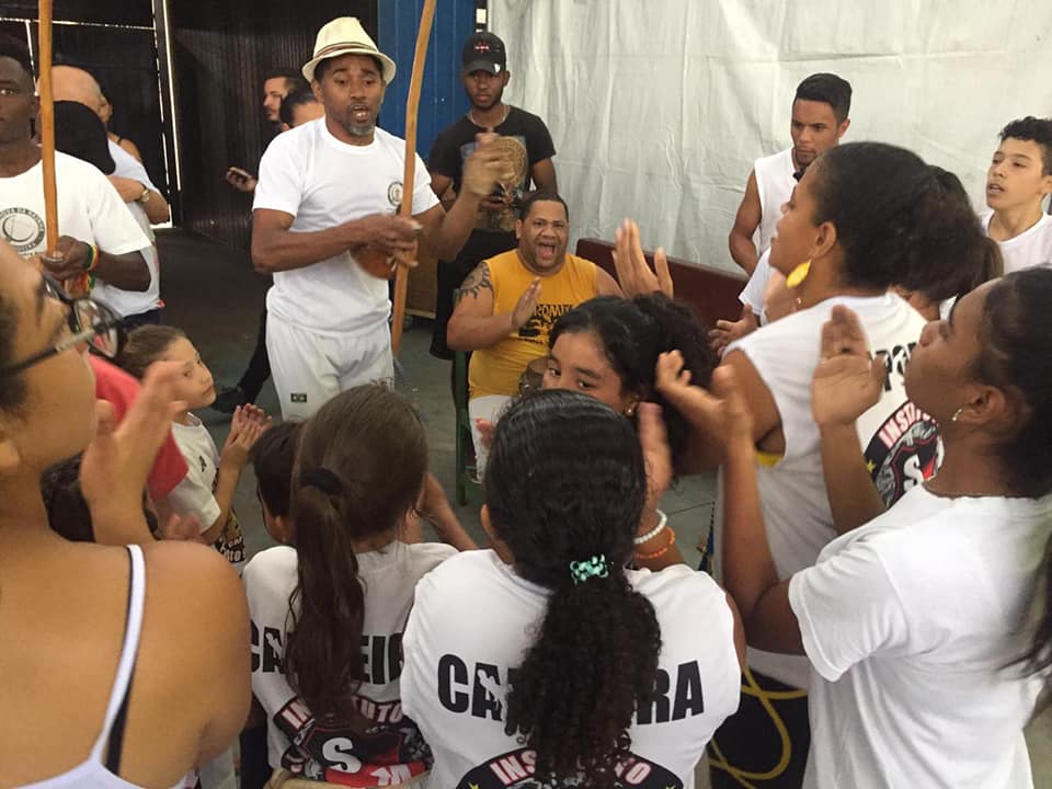 Grupo de capoeira, canta, com instrumentos típicos, no meio de uma roda de crianças e adolescentes