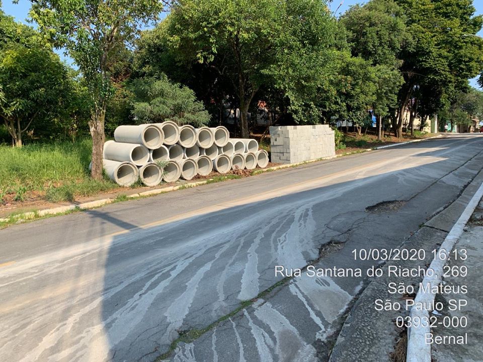 Uma pilha de tubos de cimento e outra de blocos à esquerda, à beira do asfalto, na área de uma praça.