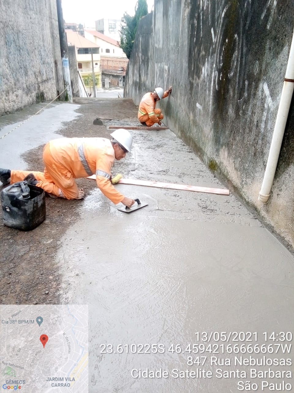 Dois homens alisam piso de uma calçada, que está sendo construída ao lado de um alambrado. 