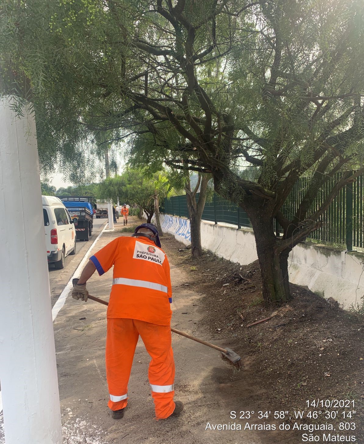 Funcionário da prefeitura, com uniforme laranja, usando ferramenta manual, recolhe detritos em uma calçada, à beira de uma mureta, com grade metálica acima. 