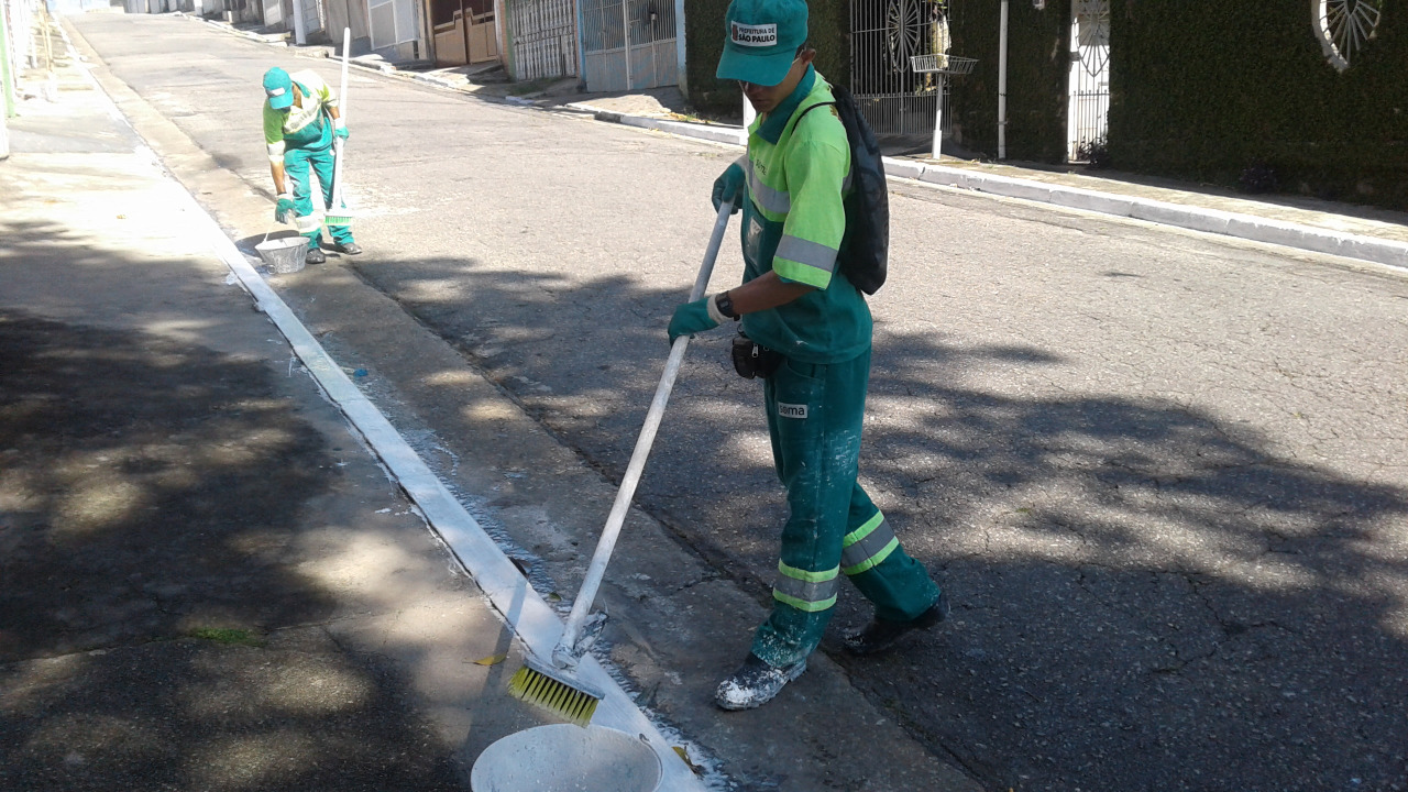 Visualiza-se dois funcionários, vestindo uniformes verdes, pintando, de branco, a guia de uma calçada.