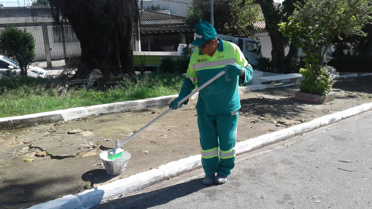 Visualiza-se um funcionário, vestindo um uniforme verde, pintando, de branco, a guia de uma calçada.