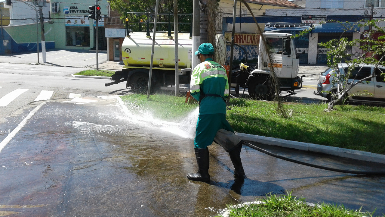 Visualiza-se um funcionário, vestindo um uniforme da cor verde, segurando uma mangueira enquanto esguicha água a fim de realizar a limpeza da rua.