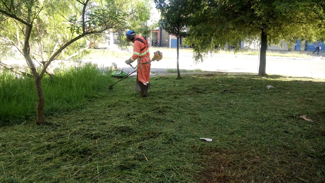 Visualiza-se um funcionário, vestindo um uniforme da cor laranja, segurando um instrumento de capinação em suas mãos enquanto realiza o serviço de corte de mato em uma praça.
