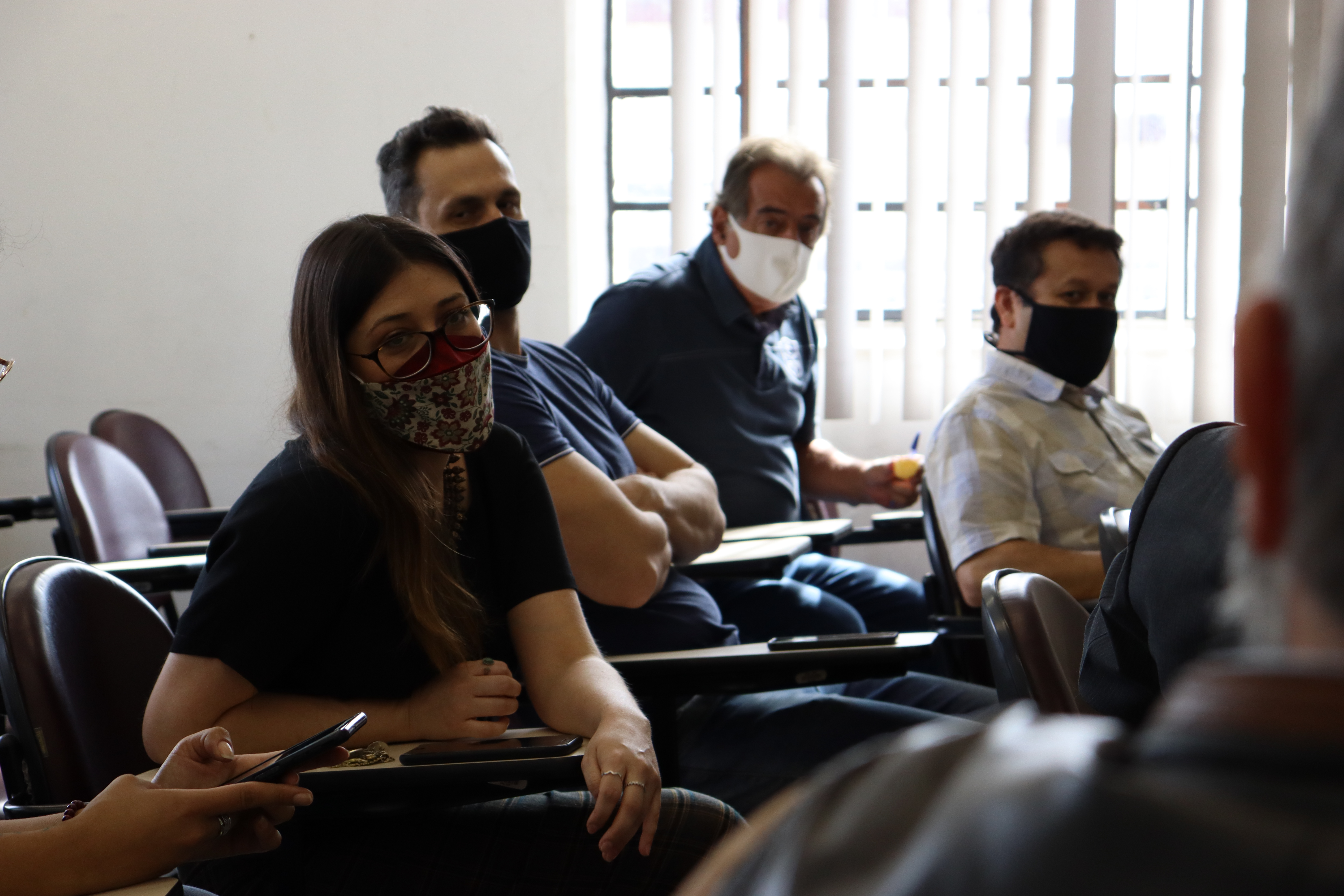 Mulher e homens sentados no auditório com máscaras nos rostos 