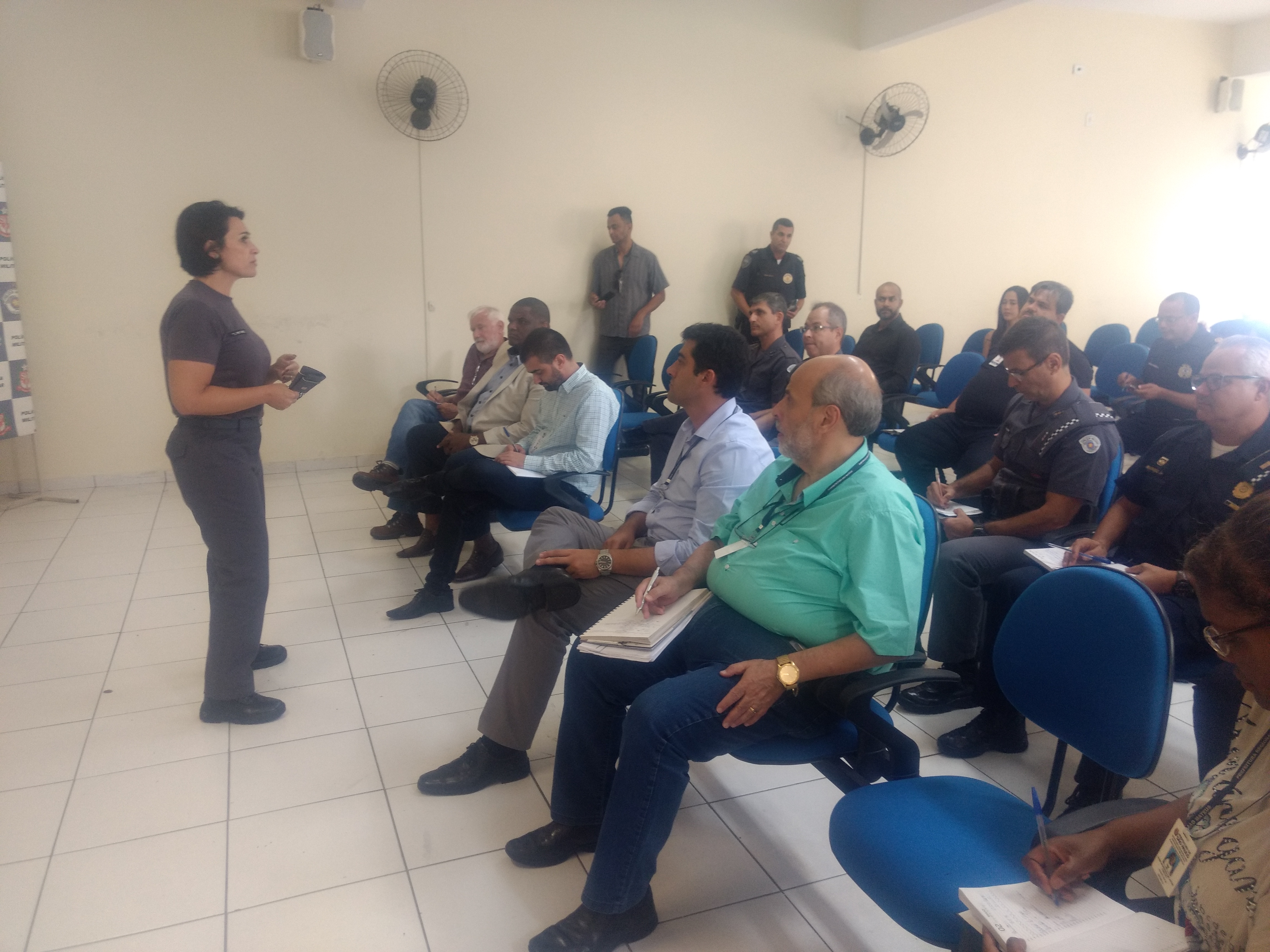 Coronel Daniele, veste a farda cinza da PM, está diante de várias pessoas, representantes de subprefeituras da zona Leste, que ouvem o que ela fala. Alguns homens estão em trajes militares 